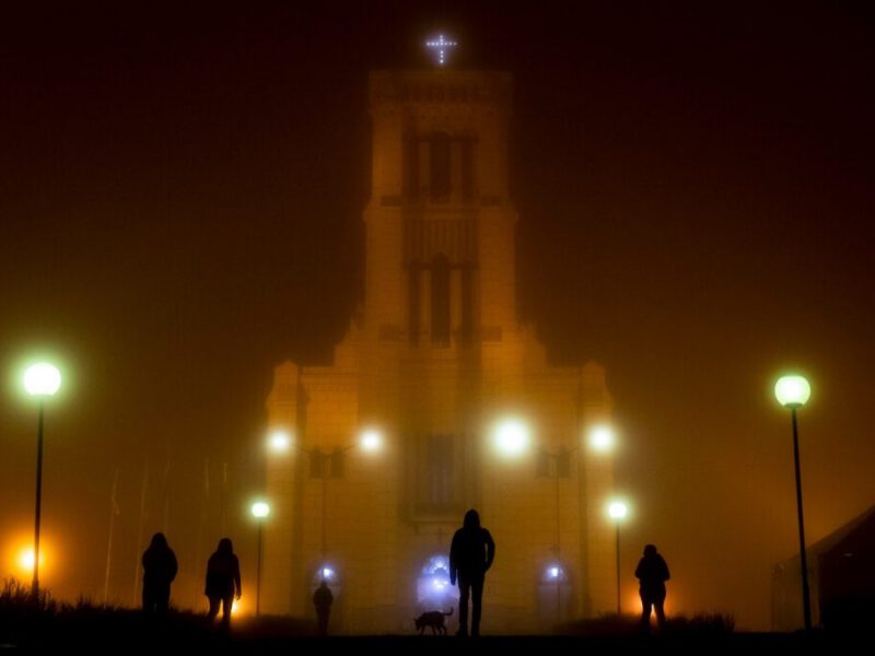 Fotografe o Jubileu: igreja iluminada na neblina noturna com fiéis em silhueta