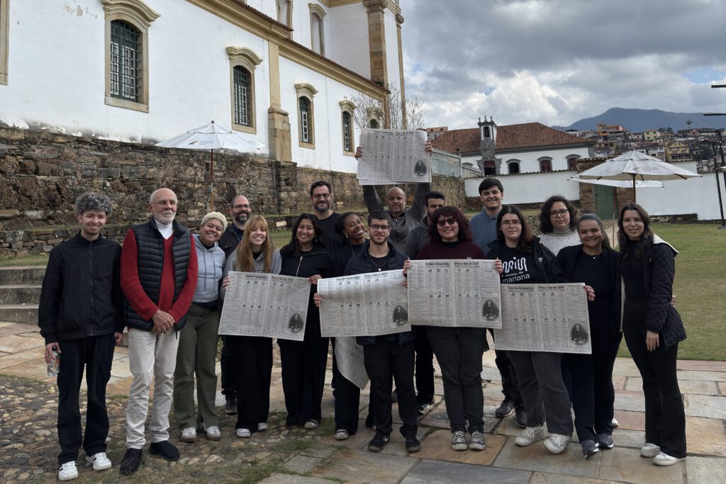 Equipe do filme Tempo Vário segura páginas da Folhinha diante de igreja em Mariana (MG)
