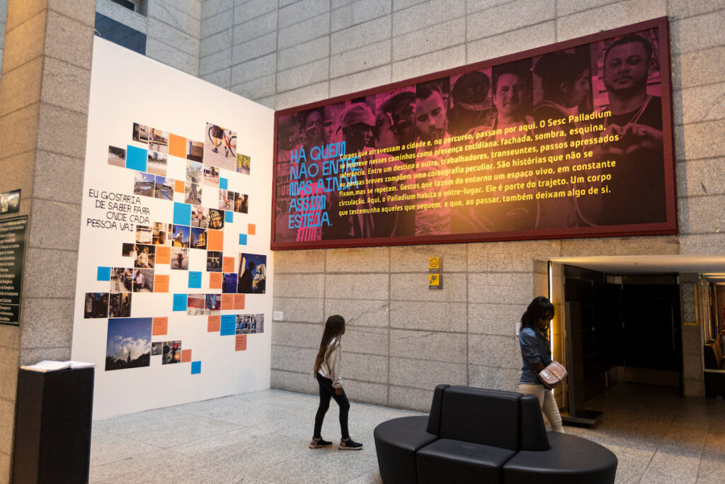 Exposição ENTRE: visitantes no saguão do Sesc Palladium diante de painéis com fotos e textos
