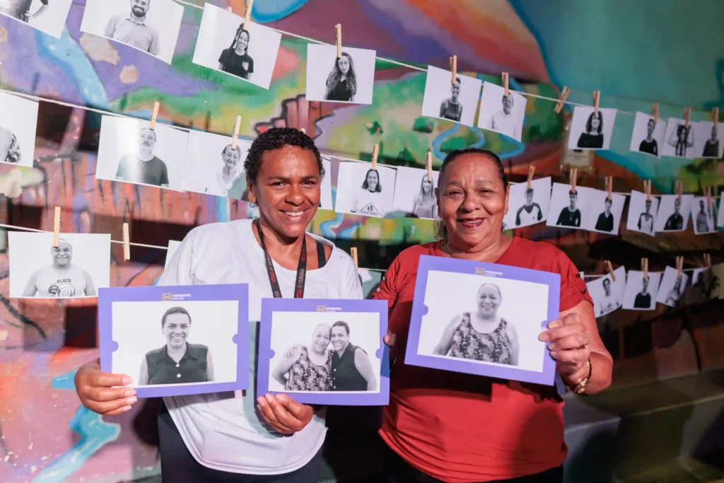 Moradores Educação: duas mulheres seguram seus retratos diante de varal de fotos