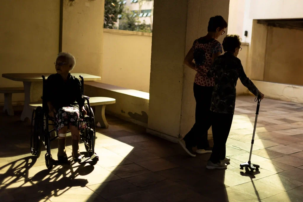 Centenários: idosas acompanhadas por cuidadora em corredor ensolarado