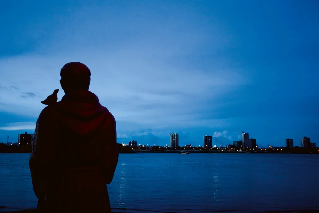 Os Chicos: silhueta de homem com pássaro no ombro diante do rio e da cidade ao anoitecer