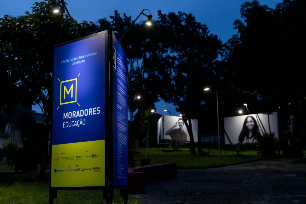 Moradores Educação: totem do projeto e retratos expostos à noite