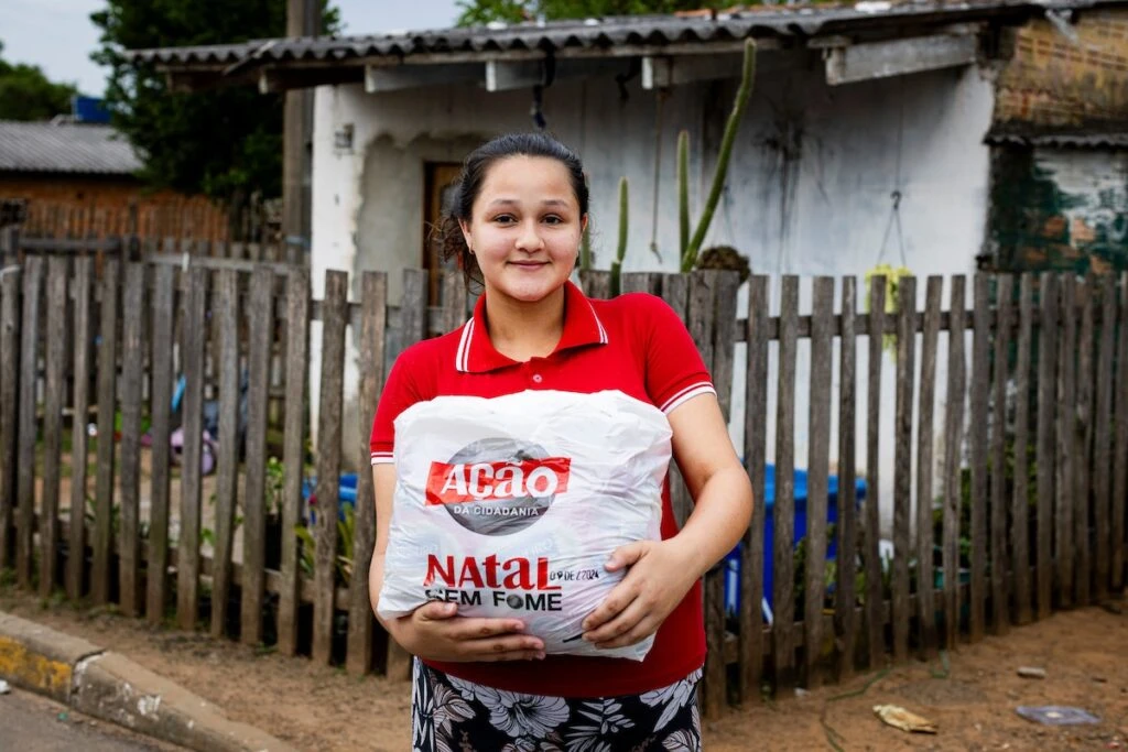 Ação da Cidadania, Natal Sem Fome: jovem sorridente segura sacola de doação diante de cerca de madeira