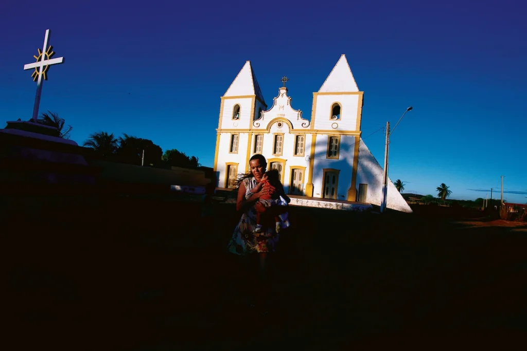 Os Chicos: mulher diante de igreja branca de duas torres sob céu azul