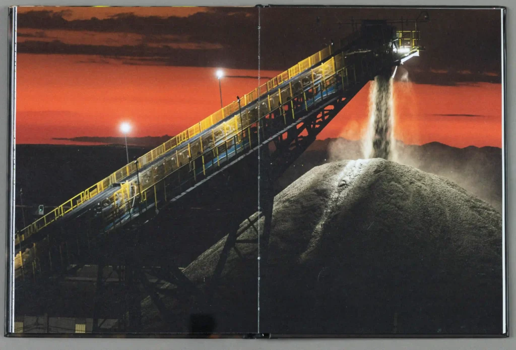 Livro Somos Todos Mineradores aberto em foto de esteira de minério ao pôr do sol
