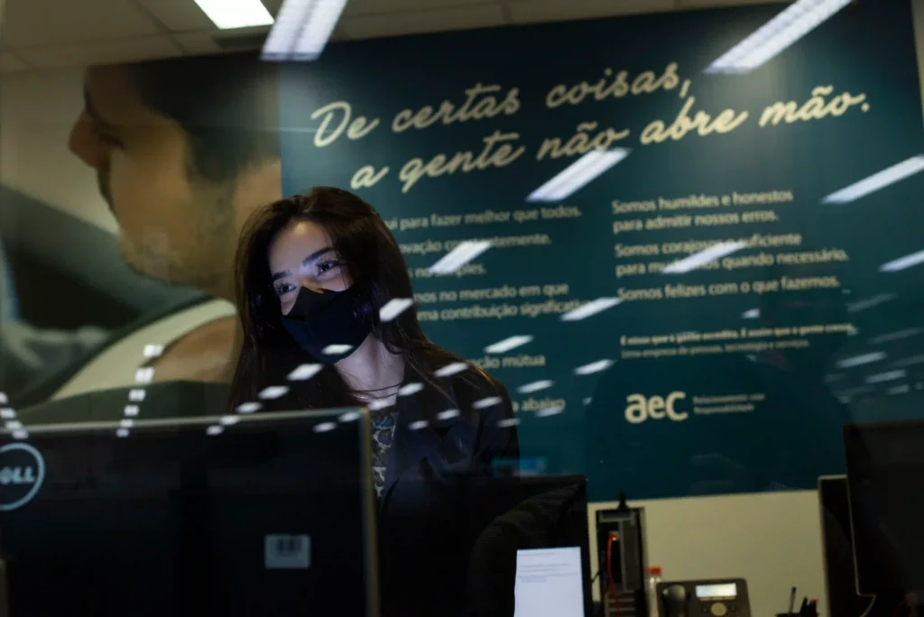 AEC: atendente de máscara diante de parede com frase da empresa