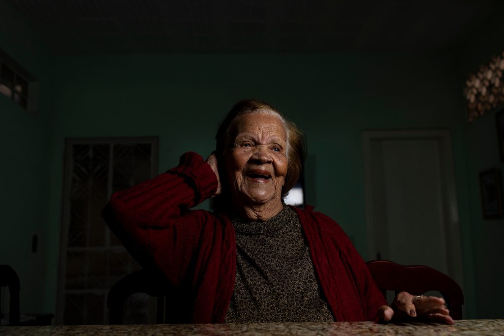 Centenários: Dona Lia, de 101 anos, sorri sentada à mesa de casa, de casaco vermelho