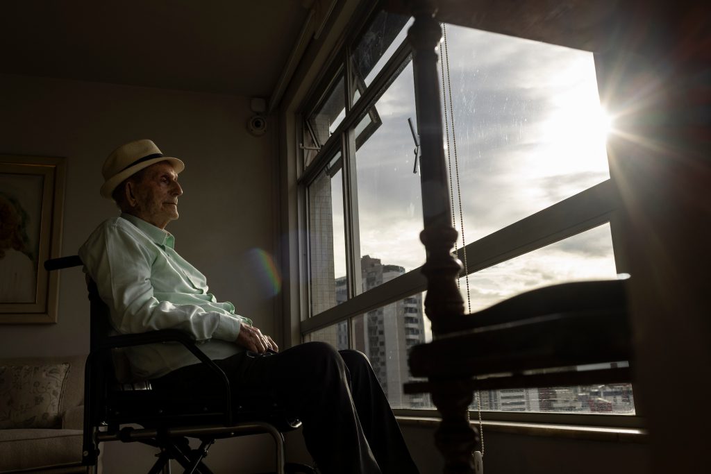 Centenários: Newton Lima, de 102 anos, de chapéu e cadeira de rodas junto à janela do apartamento